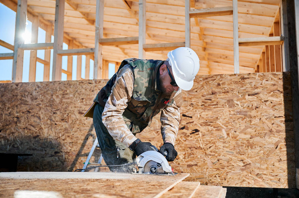 Carpenter using circular saw for cutting wooden OSB board. Man worker building wooden frame house on pile foundation. Carpentry concept. - Valokuva, kuva