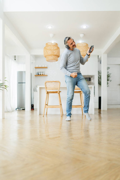 Вертикальна фотографія веселого зрілого чоловіка середнього віку в навушниках з смартфонами, що танцюють на кухні вдома
. - Фото, зображення