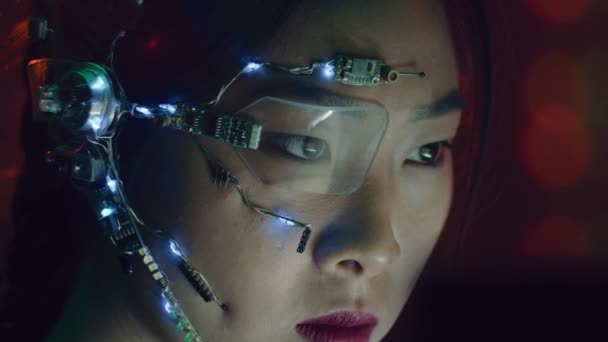 Focused asian girl in cyberpunk attire works on the computer. Wearing one-eyed glasses with white LED lights and a microphone. Neon lights background. science fiction, cybernetics concept. - Footage, Video