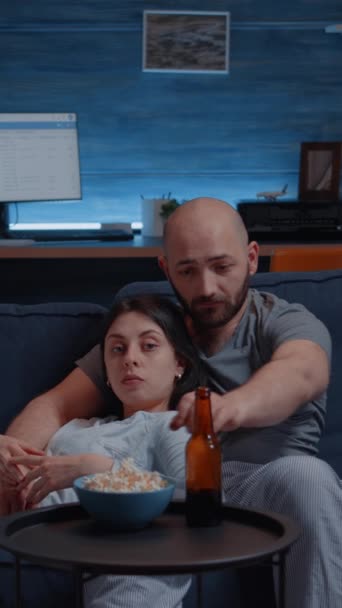 Függőleges videó: boldog pár otthon pihentető kanapén néz kedvenc TV-műsorok és eszik popcorn sörözés élvezi időt együtt otthon szabadidő, boldogság és házas emberek koncepció. - Felvétel, videó