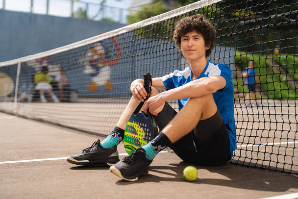 Πορτρέτο του αθλητικός νεαρός άνδρας με σγουρά μαλλιά ποζάρουν στο padel υπαίθριο γήπεδο με ρακέτα και μια μπάλα. Έννοια κηλίδες, υγιεινό τρόπο ζωής - Φωτογραφία, εικόνα