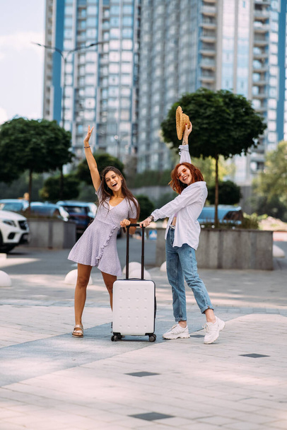 Two young women with a suitcase pose for the camera on the street - Фото, изображение