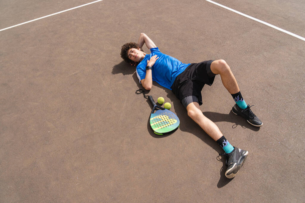 Портрет спортивного молодого чоловіка з кучерявим волоссям, що лежить на килимку на відкритому майданчику з ракеткою і м'ячем. Плями концепції, здоровий спосіб життя. Копіювати простір на землі
. - Фото, зображення