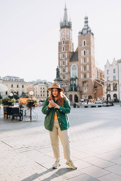 Beautiful stylish woman walking on Market Square in Krakow on autumn day and holding mobile phone. Phone Communication. Urban lifestyle concept. Check social networks - Φωτογραφία, εικόνα