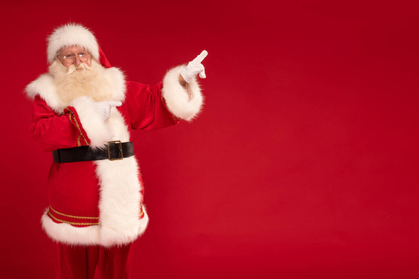 Happy bearded Santa Claus pointing on empty red studio background. Merry Christmas and Happy New Year! A lot of copy space. Xmas sale, discount concept - Foto, Imagen