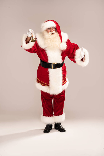 Christmas are coming! Real Santa Claus ringing the bell, standing on the light gray studio background, looking at the camera. Xmas sale, discount concept. - Photo, Image