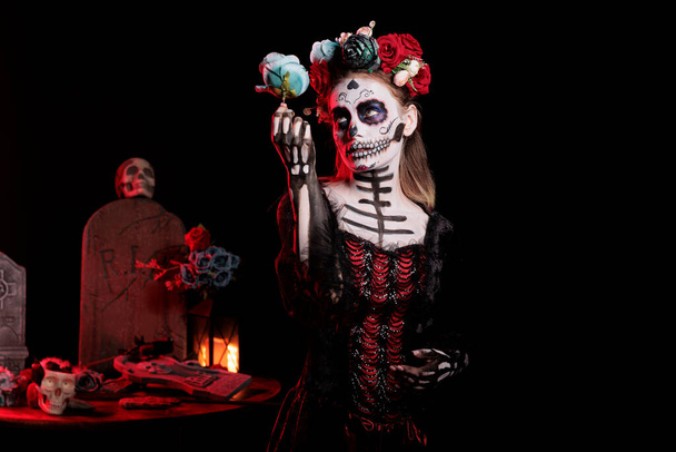 Spooky entity of death looking like santa muerte in studio, posing with roses and wearing glamour costume. Female goddess acting horror and celebrating mexican dios de los muertos holiday. - Photo, Image