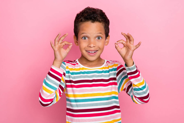 Foto de lindo chico positivo con el pelo moreno vestido a rayas manga larga mostrando signo okey aislado en el fondo de color rosa. - Foto, Imagen