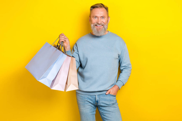 Photo of cheerful aged person arm hold boutique bags toothy smile isolated on yellow color background. - Foto, Bild