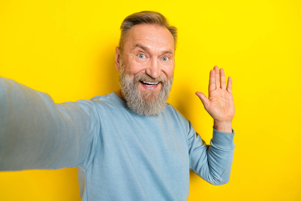 Photo of attractive friendly retired guy wear blue sweater tacking selfie waving arm palm isolated yellow color background. - Foto, Imagem