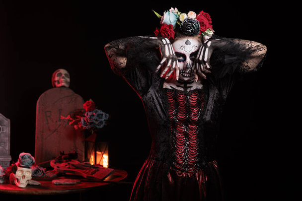 Piękna kobieta zachowuje się jak bogini śmierci z tradycyjną sztuką ciała czaszki i koroną kwiatów, wyglądająca jak święty Mikołaj muerte w dzień śmierci. Czarująca osoba pozująca do meksykańskiej tradycji.. - Zdjęcie, obraz
