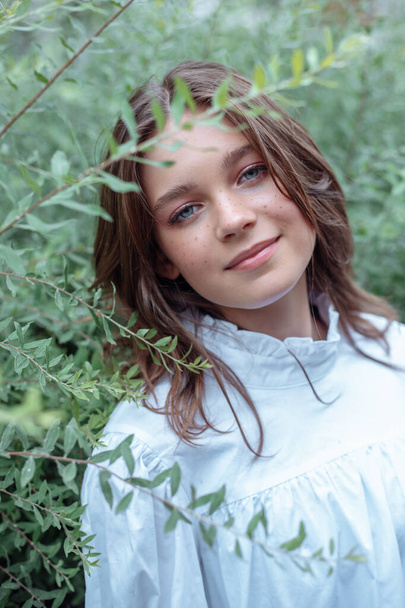 Portrait closeup of cute young girl in nice white dress with smile standing among branches of bushes with green leaves, nature background. Walking in summer city park, forest, natural beauty. - Photo, image