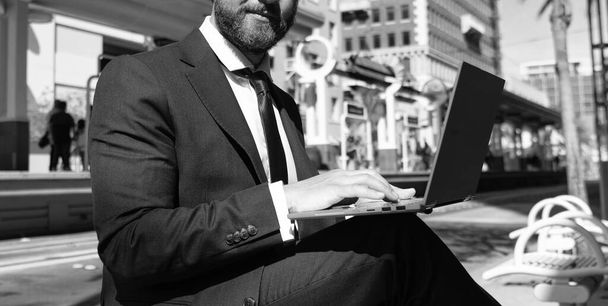 Kesilmiş takım elbiseli işadamı kentteki bankta oturuyor dizüstü bilgisayar ve iş bloglarında çalışıyor.. - Fotoğraf, Görsel
