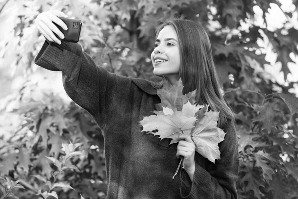 秋の自然。美しい顔の肖像画。自然の美しさ。秋のファッション。秋の葉で自撮りしてる女の子。幸せな女性はスマートフォンを持ち. - 写真・画像
