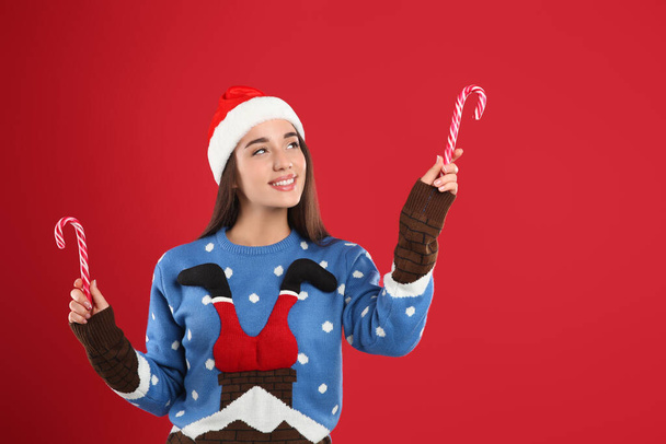 Young woman in Christmas sweater and Santa hat holding candy canes on red background - Foto, afbeelding