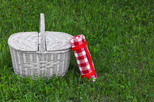 Rolled checkered tablecloth near picnic basket on green grass outdoors, space for text - Фото, изображение