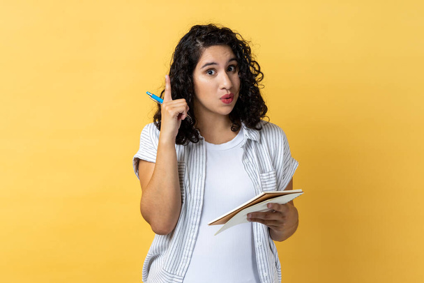 Portrait of amazed inspired woman journalist with dark wavy hair writing in paper notebook, having new excellent idea, raising finger up. Indoor studio shot isolated on yellow background. - Foto, Bild