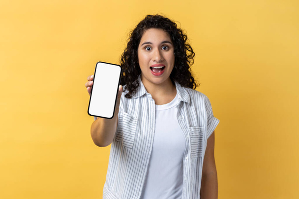 Portrait of excited amazed woman with dark wavy hair holding phone with empty display, advertisement area, copy space for promotional text. Indoor studio shot isolated on yellow background. - Photo, image