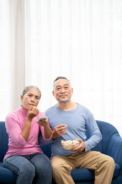 Glücklich lächelndes asiatisches Seniorenpaar, das auf dem Sofa sitzt und zu Hause im Wohnzimmer fernsieht. Senioren verbringen Wochenende zusammen. - Foto, Bild