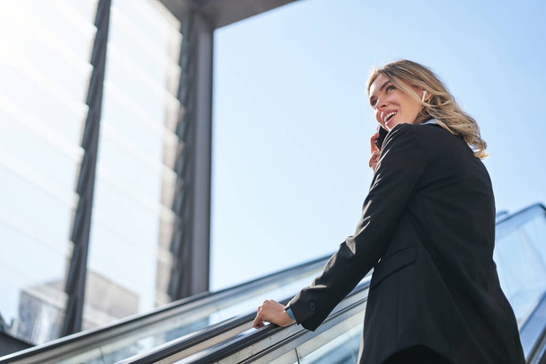 Portrait of businesswoman in black suit, going up on escalator, talking on mobile phone. Saleswoman walking in city, having a call. - Photo, Image