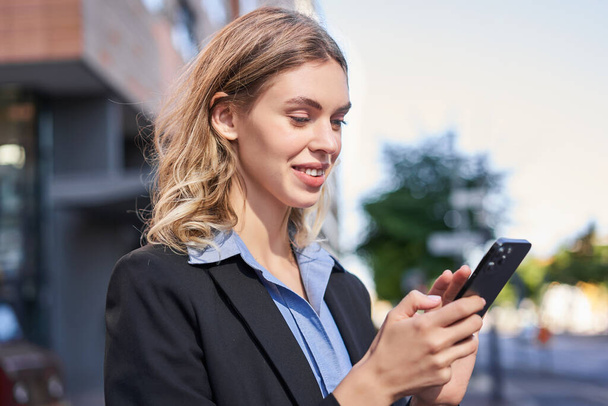 Donna aziendale si trova sulla strada e messaggi di testo sul telefono cellulare, sorridendo guardando lo schermo dello smartphone. - Foto, immagini