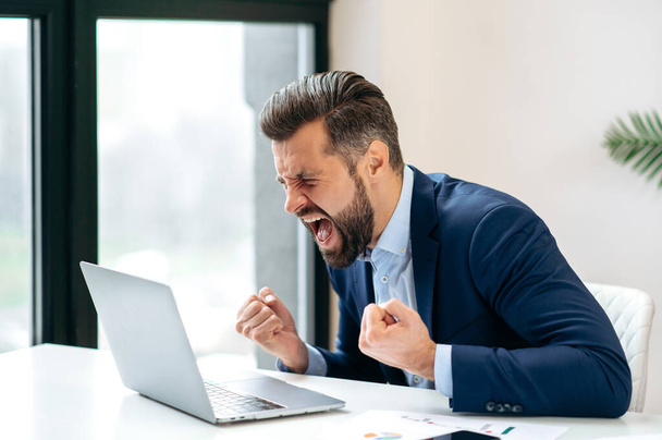 Angry annoyed frustrated caucasian business man, sitting at workplace with laptop, screaming, gesturing with fists, failed deal, disappointed with project result, experiencing negative emotions - Foto, Imagen