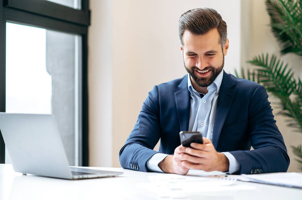 On-line zprávy. Pozitivně obsazeno kavkazský obchodník v obleku, zaměstnanec společnosti, top manažer, sedí v kanceláři, pomocí svého chytrého telefonu, smskuje online s kolegou nebo klientem, usmívá - Fotografie, Obrázek