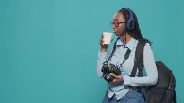 Woman listening to music on headset and having coffee cup, travelling on holiday vacation to take pictures as professional ptohographer. Carrying backpack and camera, enjoying song. - Video