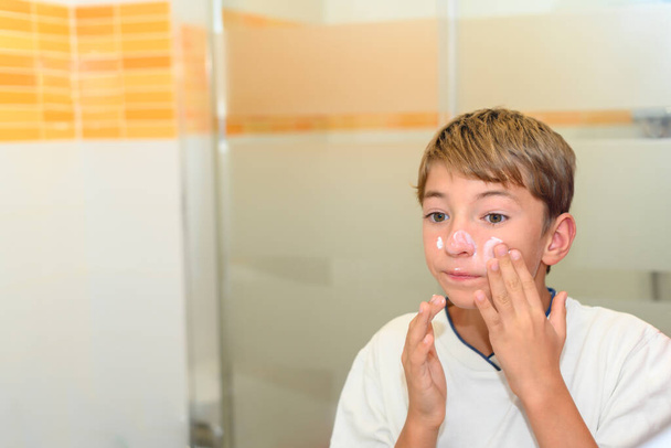 A preteen puts moisturizer on her face with her hands in front of the bathroom mirror. - Fotoğraf, Görsel