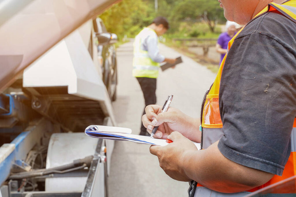 Closeup and crop insurance agent writing on clipboard while examining car after accident claim being assessed and processed on blurred damaged car truck slides with peoples and sun flare background. - Zdjęcie, obraz