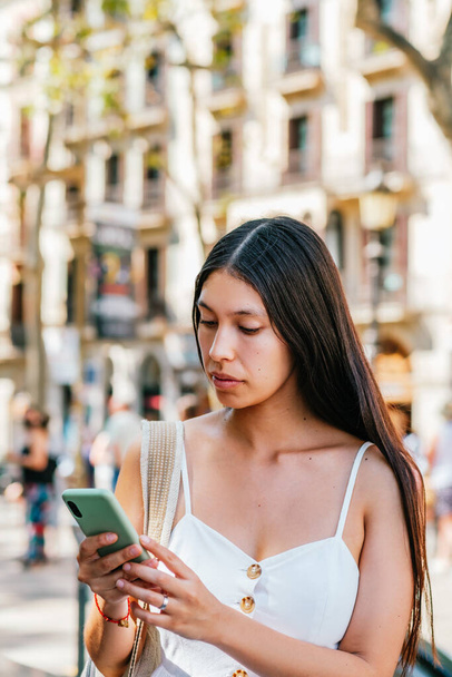 スペインのバルセロナで晴れた夏の日に路上のぼやけた背景に立っている間、スマートフォン上のソーシャルメディア上でラテンアメリカの女性のスクロールニュース - 写真・画像
