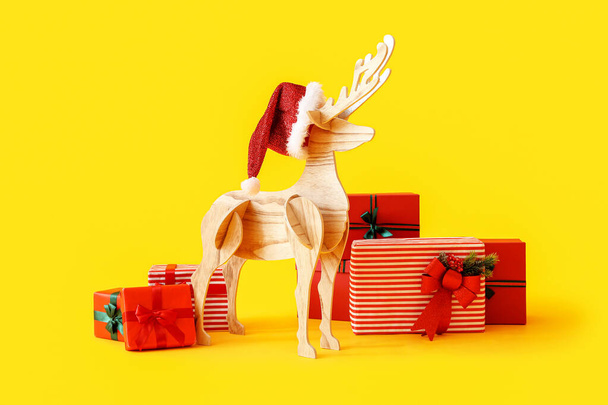 Wooden reindeer with Santa hat and Christmas presents on yellow background - Фото, изображение