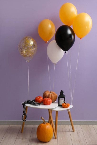 Mesa con calabazas, cadena, linterna y globos de Halloween cerca de la pared violeta - Foto, Imagen