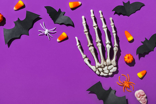 Frame made of Halloween treats, paper bats and skeleton hand on purple background - Фото, изображение