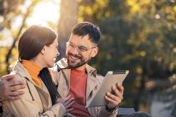 公園のベンチに座って楽しんでいる若いカップルは、デジタルタブレットを使用して. - 写真・画像