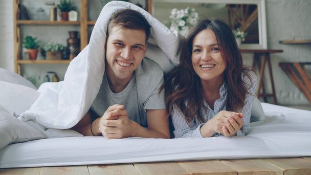 Portrait of happy couple lying in bed under blanket looking at camera laughing and smiling. Loving married people and happiness concept. - Foto, Bild