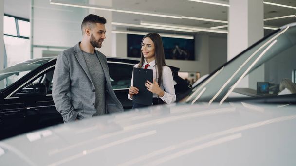 Attractive female car dealer is talking to interested customer about new automobile model and holding documents while standing together near beautiful expensive car. - Photo, Image