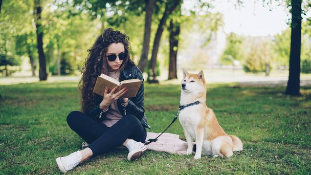 Attractive young woman is reading book sitting on grass in park while her well-bred dog is sitting near her owner and looking around. Leisure, nature and animals concept. - Photo, Image