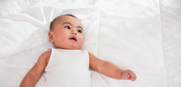Asian smiling infant child at home, Cute little baby on bed under soft blanket, baby wearing bodysuit lying on white beedsheets in bedroom - Photo, Image