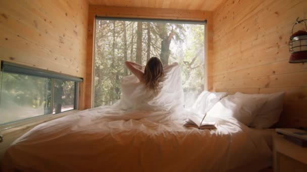 Задний вид на красивую голую лесную фею с длинными волнистыми волосами в таинственном туманном горном лесу солнечным утром с солнечными лучами, сияющими в деревянной уютной каюте на кровати. Волшебный мир в 4K - Кадры, видео