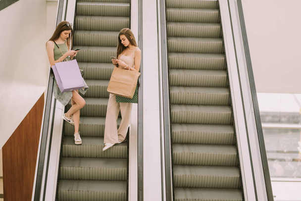 Dwie słodkie dziewczyny patrząc na swoje telefony podczas schodzenia na schodach ruchomych w centrum handlowym - Zdjęcie, obraz