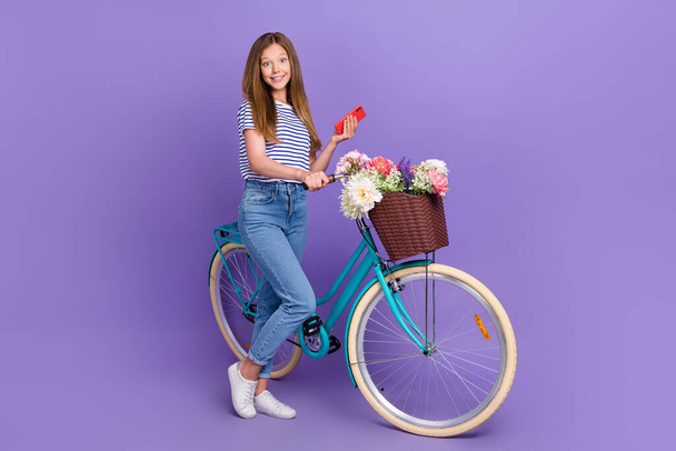 Full length photo of funny sweet small schoolgirl wear striped t-shirt typing modern device riding bicycle isolated purple color background. - Photo, image