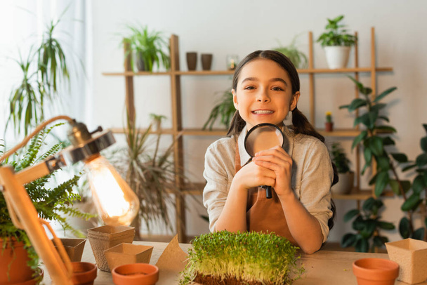 Smiling kid holding magnifying glass near blurred plants and lamp at home  - Fotó, kép