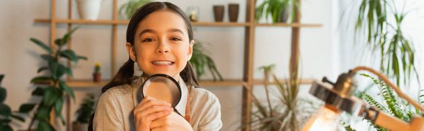 Cheerful girl holding magnifying glass and looking at camera near blurred lamp at home, banner  - Photo, Image