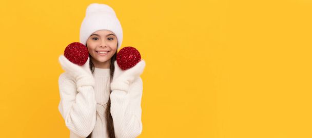 cheerful child in winter hat and gloves hold decorative xmas balls on yellow background, xmas. Banner of christmas child girl, studio kid winter portrait with copy space - Фото, изображение