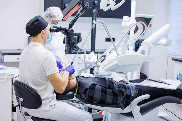 Стоматолог и ассистент проверяют зубы пациента с помощью стоматологических инструментов - микроскопа, зеркала и зонда в кабинете стоматологической клиники. Концепция медицины, стоматологии и здравоохранения. Стоматологическое оборудование - Фото, изображение