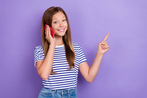 Photo of optimistic girl with straight hairdo wear striped t-shirt talk on phone directing empty space isolated on purple color background. - Foto, Bild