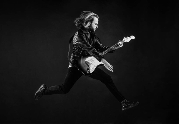 musician guitar player jumping. playing rock music. stylish crazy man. string musical instrument. emotional bearded rocker in leather jacket. man long hair play electric guitar. rock music style. - Foto, afbeelding