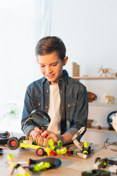 Smiling boy holding magnifying glass near blurred robotic model at home  - Photo, Image