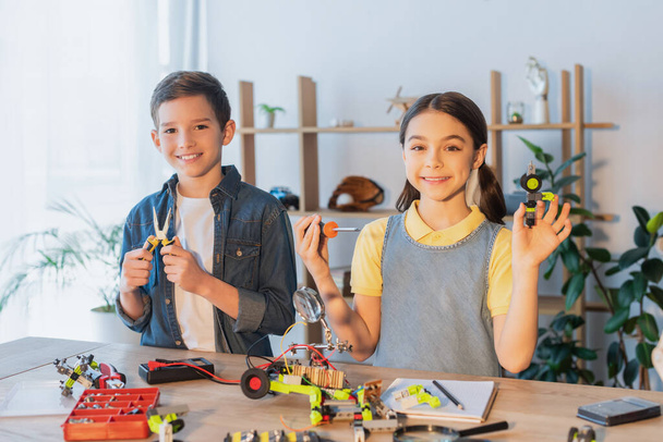 Crianças felizes segurando ferramentas perto do modelo robótico na mesa em casa  - Foto, Imagem